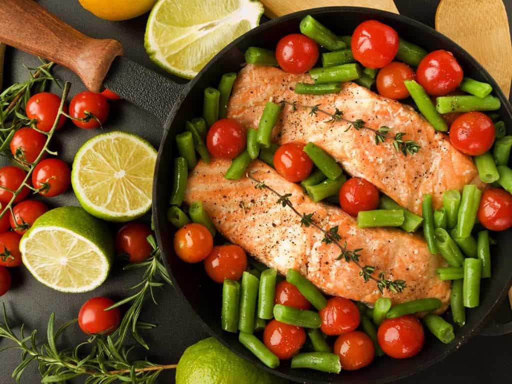 salmon and vegetables in a cast iron skillet, black countertop and sliced limes outside of pan