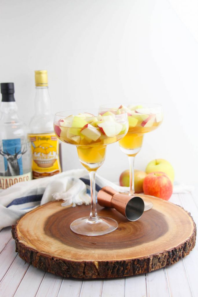 Apple Cider Margarita, two of them on the round wooden serving tray with ingredients show in the background as well. 