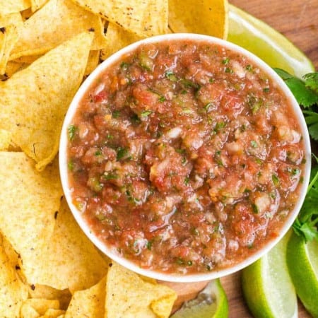 tortilla chips on left, fresh cilantro and lime wedges on the right with white bowl of salsa in the middle