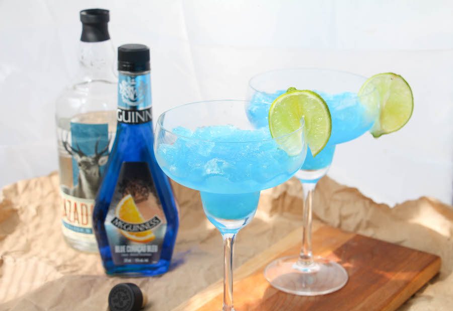 Two frozen blue margaritas on a wooden board with ingredients in the back. 