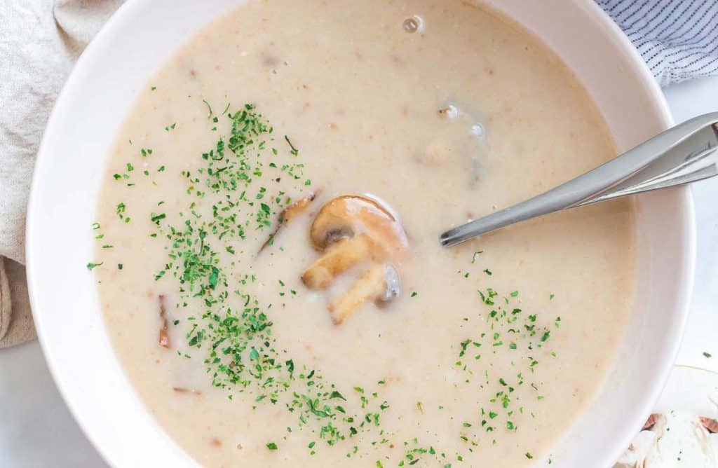 close up of a white bowl filled with cream of mushroom soup. THere is parsley sprinkled on the bowl of soup and a mushroom is visible under the broth. 