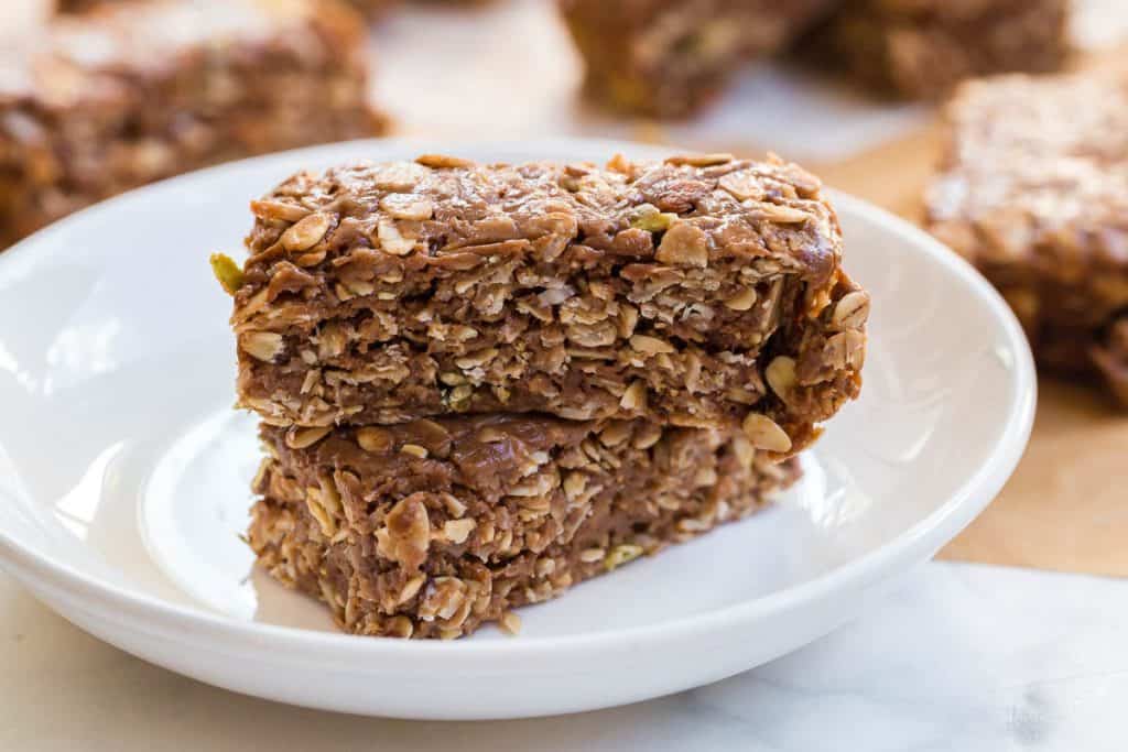 two homemade peanut butter granola bars on a white plate. in the background you can see more granola bars on butcher paper.