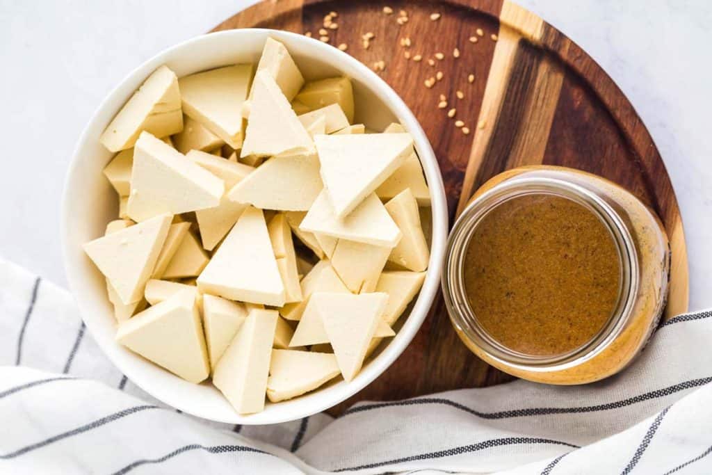 image of raw tofu cut into triangles in a white bowl with a small clear mason jar of homemade teriyaki sauce. Wood charger and white striped napkin in the background. 