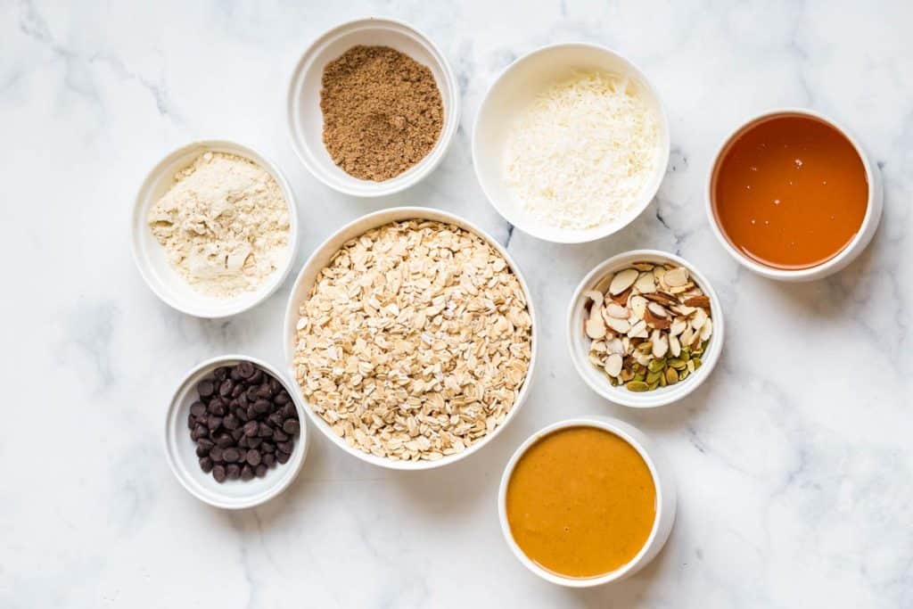 ingredients for peanut butter granola bars in white bowls on top of a white marble countertop