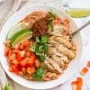 white counter with white bowl filled with cauliflower rice, shredded chicken thighs, and fresh vegetables