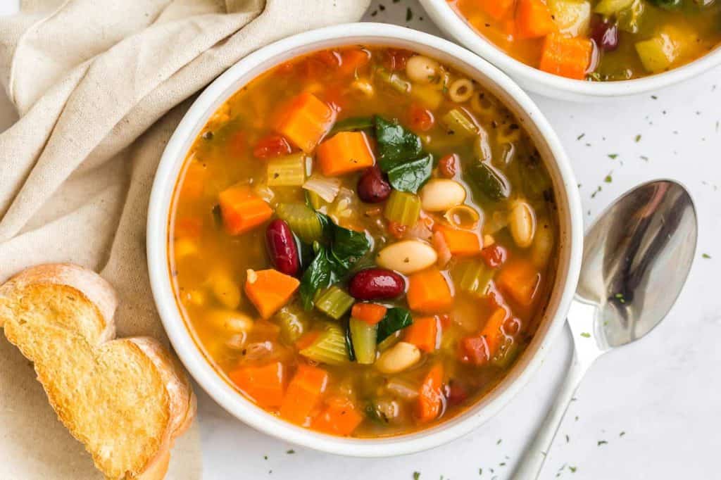 cooked minestrone soup in white bowl with crusty bread and napkin on counter