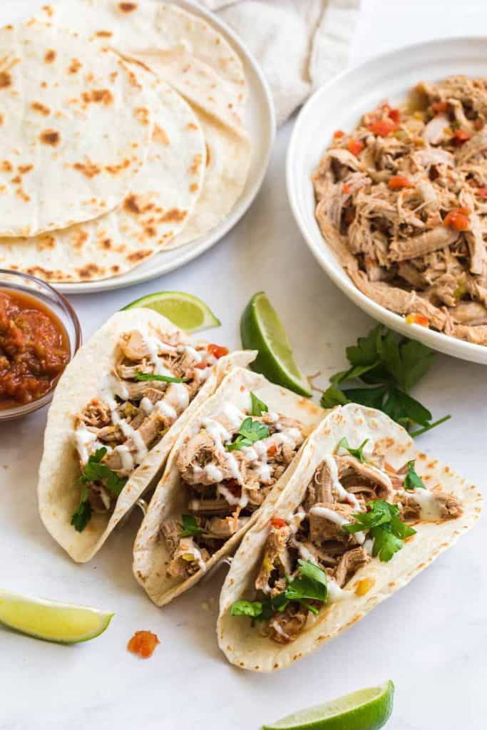 three pork carnitas tacos with flour tortillas, bowl of carnitas nearby