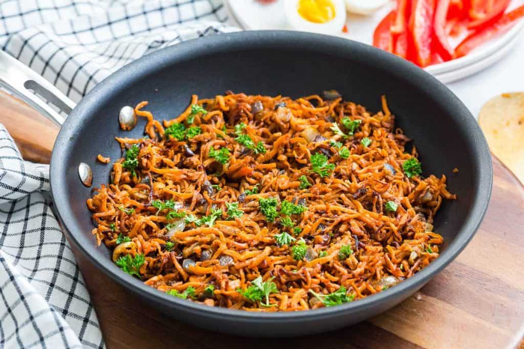 black pan with cooked sweet potato hash browns and onions with decorative parsley on top. In the background is a wood tray, blue and white napkin, and a white plate with hard boiled eggs, bacon, and sliced red peppers. 