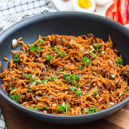 black pan with cooked sweet potato hash browns and onions with decorative parsley on top. In the background is a wood tray, blue and white napkin, and a white plate with hard boiled eggs, bacon, and sliced red peppers.