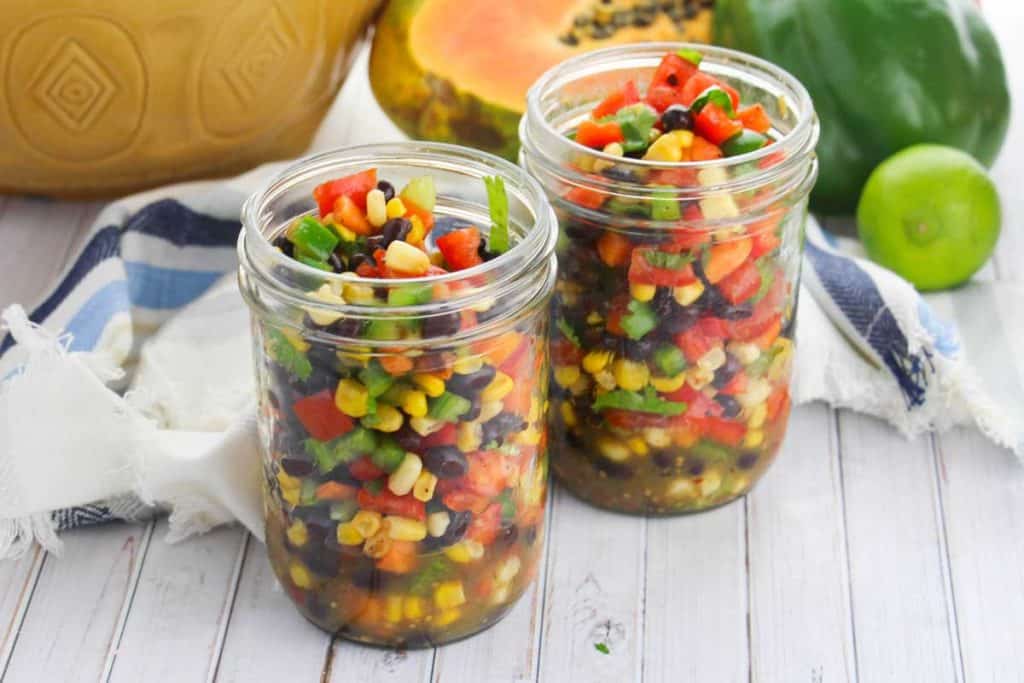 two small mason jars full of summer confetti salad with fresh food in the background