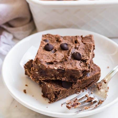 horizontal image of two black bean brownies stacked on a white plate with a fork next to them. Countertop is white, as is a pan of brownies in the background.