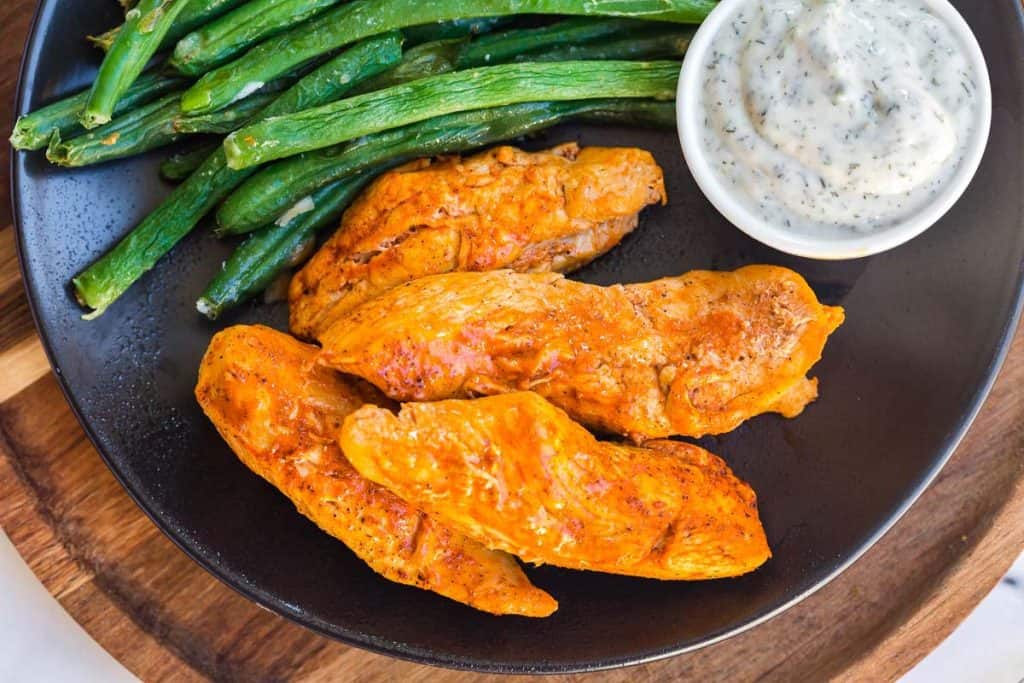 black plate with cooked boneless buffalo wings and green beans, with small white dish of ranch dressing on the side.